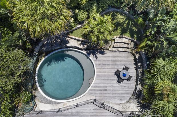 Aerial view of a landscaped outdoor pool area with a rounded, organically shaped swimming pool beside a broad weathered timber deck. Dense tropical planting surrounds the space, with steps, stone or concrete edging, and dark metal railings visible around the perimeter. A small outdoor table and chairs sit on the deck.