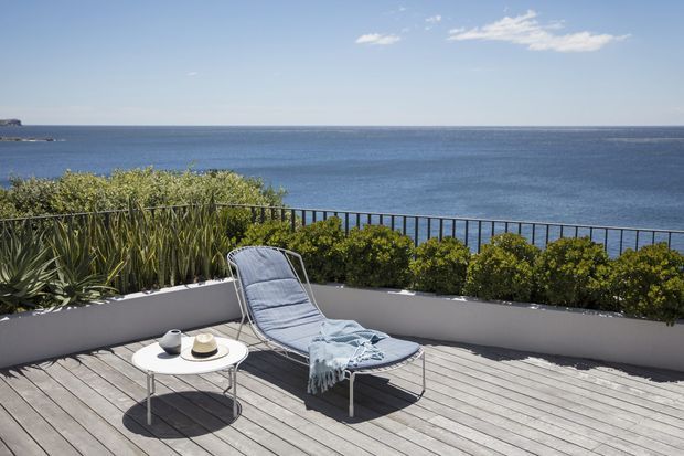 Sunny coastal terrace overlooking the ocean, furnished with a reclining lounge chair and small round side table on a weathered timber deck. A raised white planter bed with coastal planting and a dark metal balustrade frame the expansive sea view.