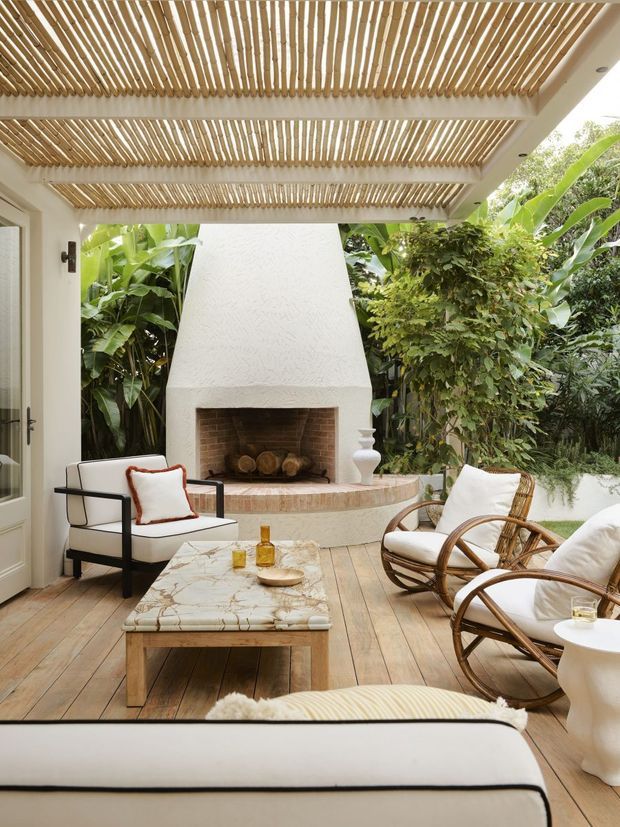 A bright covered outdoor lounge or patio with a timber slatted pergola overhead, warm timber decking, a white rendered masonry fireplace with brick-lined firebox, and a curved tiled hearth. The space is furnished with white upholstered seating, rattan lounge chairs, and a marble-topped timber coffee table, surrounded by lush tropical planting.
