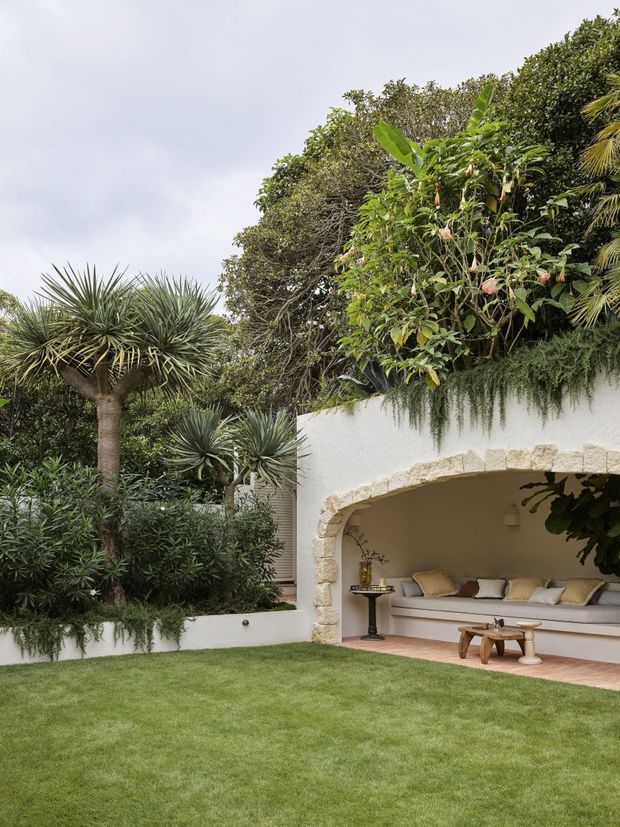 A lush landscaped courtyard garden with tropical and subtropical planting, manicured lawn, and a white rendered outdoor lounge pavilion. The pavilion features a large rounded stone-trimmed arched opening, built-in upholstered seating with neutral cushions, and a small timber coffee table. Trailing greenery softens the roofline and walls, creating a resort-like outdoor entertaining space.