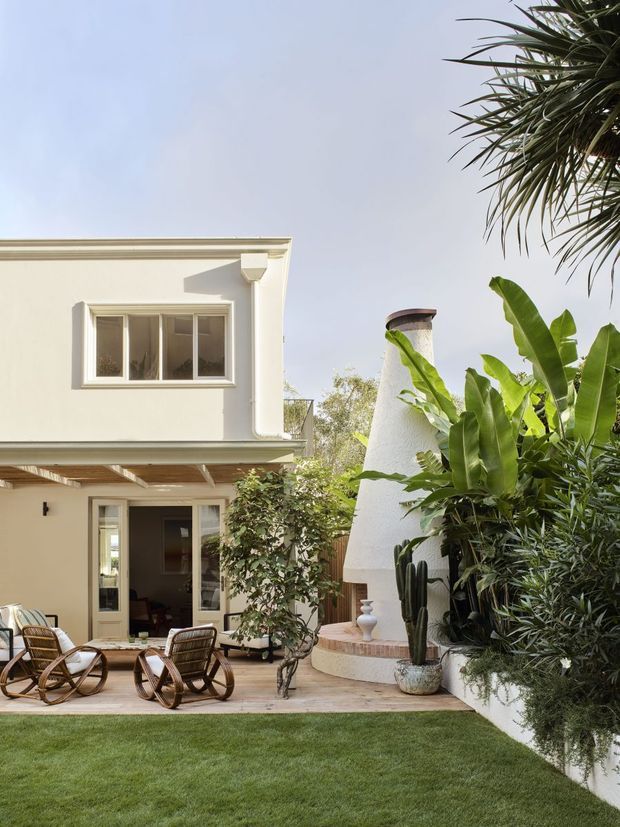 A landscaped courtyard at the rear of a two-storey cream-rendered residence. The space features a timber pergola over glazed doors, a paved patio with woven lounge chairs, a sculptural white stucco outdoor fireplace or chimney, raised curved brick-edged planting, and lush tropical greenery surrounding a lawn.