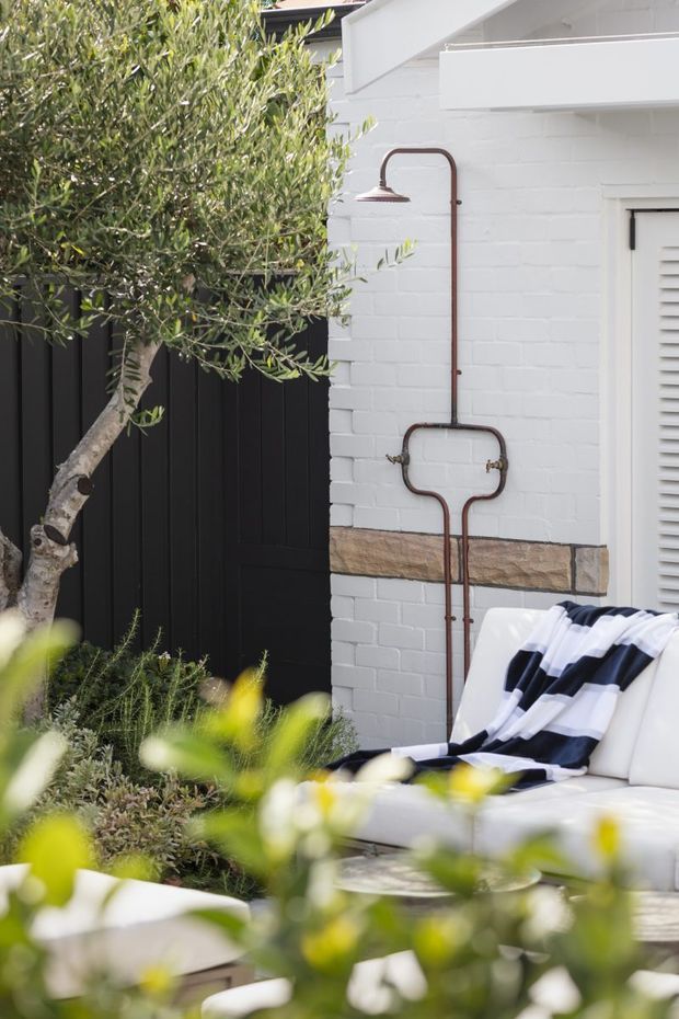 A styled outdoor courtyard or patio corner beside a white-painted brick wall, featuring a vintage copper wall-mounted outdoor shower, black vertical fencing, an olive tree, low landscaping, and a white outdoor sofa with a striped throw. The scene has a relaxed, refined garden-lounge character.