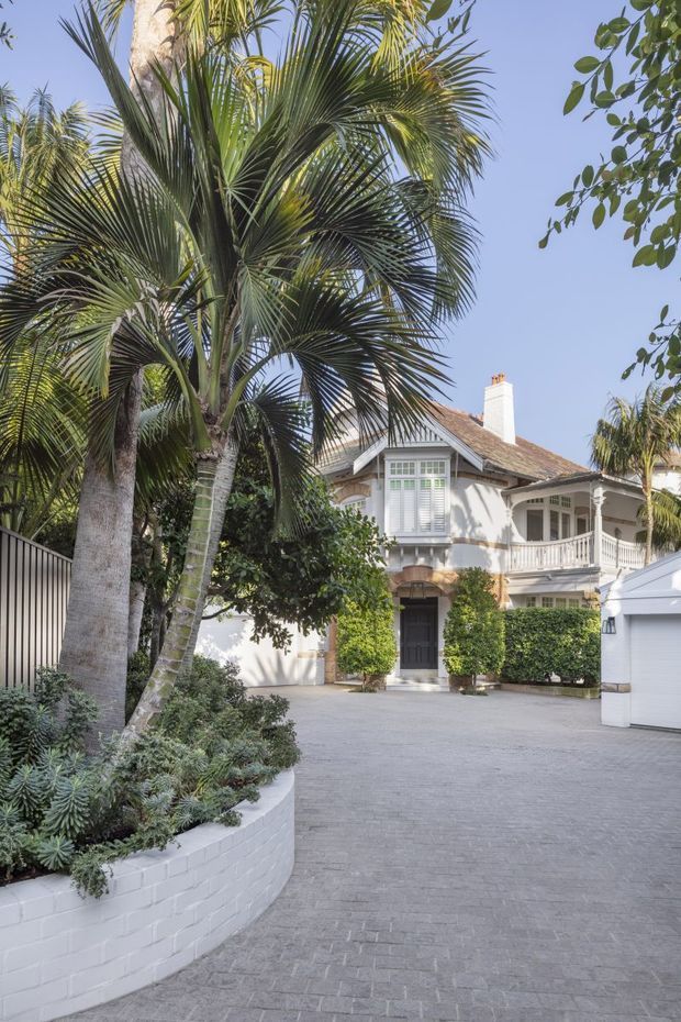 A bright exterior view of a refined two-storey residence, partially screened by mature palm trees and lush landscaping. The home features a pitched shingle roof, white rendered walls, decorative timber gable detailing, a projecting bay window with shutters, balcony railings, and a covered entry porch. A broad grey paved driveway leads toward the entrance and adjacent garage, with white-painted brick garden walls and clipped hedges framing the approach.