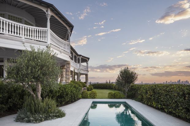 Exterior view of a grand two-storey residence with ornate white verandas, decorative balustrades, columns, and a dark pitched roof. A narrow rectangular swimming pool and pale paved terrace sit within a manicured garden, with an urban skyline visible in the distance at sunset.