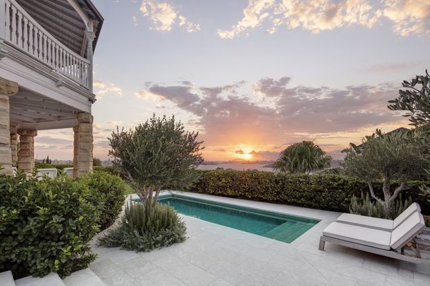A landscaped outdoor pool terrace beside a substantial coastal-style house, with pale stone paving, a rectangular swimming pool, hedges and mature planting. A covered veranda with stone columns and white balustrading occupies the left side, while the terrace overlooks a dramatic sunset and distant water or horizon.