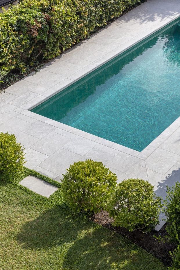 A rectangular swimming pool with clear turquoise water, surrounded by large-format light grey paving. Manicured hedges, rounded shrubs, and lawn border the pool, creating a neat landscaped outdoor setting.