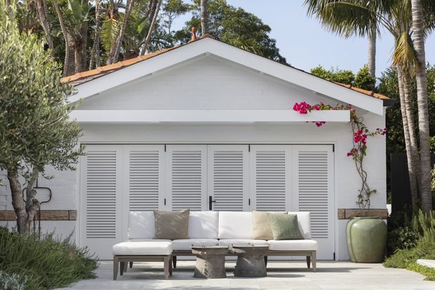 A bright tropical-style exterior courtyard or patio scene in front of a small white gabled structure. The facade features louvered white doors, a terracotta-edged pitched roof, and a simple covered overhang. A white outdoor sofa with cushions and a low coffee table sits on pale paving, framed by palms, greenery, flowering vines, and a large ceramic planter.
