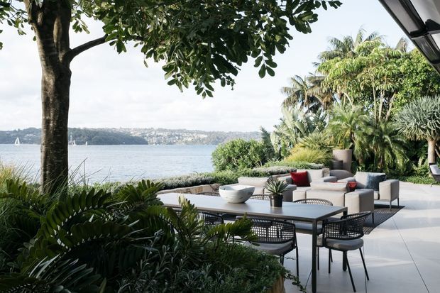 A landscaped waterfront outdoor terrace with a large dining table and woven dining chairs in the foreground, and a separate lounge seating area with neutral sofas and red accent cushions beyond. Lush tropical planting frames views across the water to a distant shoreline. The terrace has large light-coloured paving tiles and a contemporary covered edge.