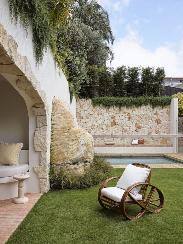 A landscaped courtyard garden with a small rectangular swimming pool, stone feature wall, white rendered arched structure, lush greenery, and a rattan lounge chair on the lawn. The space has a relaxed Mediterranean-inspired outdoor character with natural stone and abundant planting.