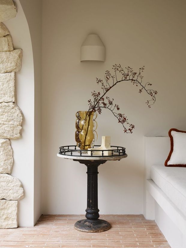 A softly lit interior vignette featuring a small round black pedestal table with a decorative open metal rim, a translucent amber glass vase, and dried branches. The table stands against a warm off-white plaster wall beneath a simple wall sconce. A rough natural-stone wall edge is visible on the left, while a built-in white upholstered bench with a rust-brown trimmed cushion appears on the right. The floor is finished in narrow terracotta-toned brick or brick-look tiles, creating a warm, textural and refined atmosphere.