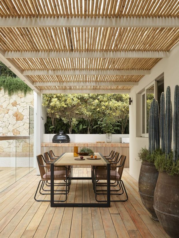 A refined outdoor dining terrace beneath a timber pergola with closely spaced bamboo or timber slats. The space features a long dining table with black metal-framed chairs, timber decking, a built-in outdoor kitchen or barbecue counter, stone wall cladding, lush planting, and large potted cacti. The partially enclosed courtyard-like setting has a warm, natural, contemporary feel.
