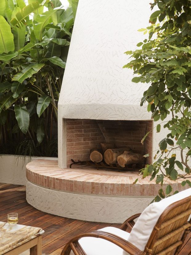 Outdoor living area featuring a large built-in fireplace with a white textured stucco surround, curved brick hearth and firewood storage. The fireplace sits beside dense tropical planting on a timber deck, with woven rattan seating visible in the foreground.