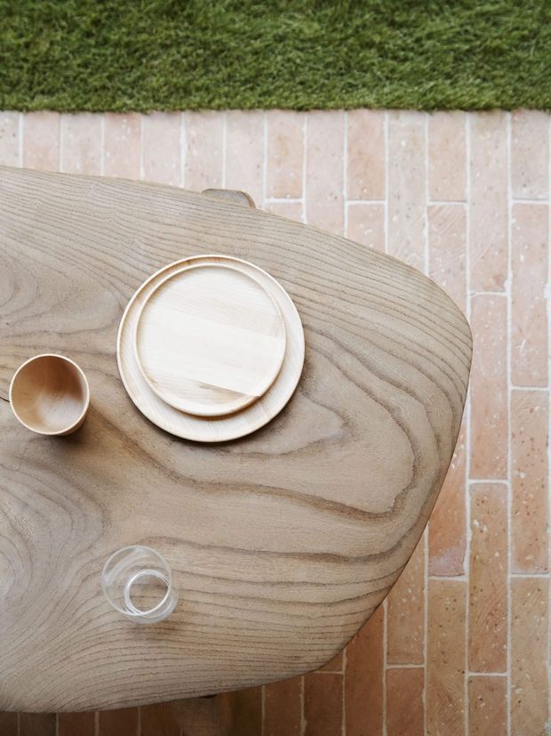 Top-down view of a softly rounded light timber dining table with visible natural grain, set with pale ceramic plates and wooden-toned tableware, a small cup, and a clear glass. The table sits on a warm, light brick-tiled floor beside a strip of green carpet or turf, creating a natural, tactile dining setting.