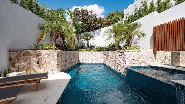 A bright, modern courtyard pool area featuring a narrow turquoise swimming pool and raised spa with dark mosaic tile, surrounded by pale irregular stone retaining walls, white rendered walls, tropical palms and planting, and timber vertical screening. Lounge furniture sits beside the pool under a clear blue sky.