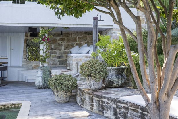 A tranquil outdoor courtyard or poolside terrace with a weathered stone wall, timber decking, white-painted architectural elements, built-in seating, potted shrubs, and mature trees. The space has a relaxed coastal character and appears to connect to a pool at the lower left.