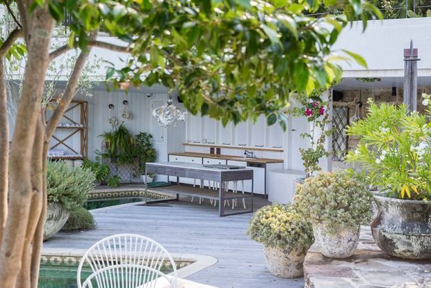 A bright outdoor entertaining area with a weathered timber deck, outdoor dining table and chairs, white built-in cabinetry, hanging pendant lights, and lush potted garden planting. The space is framed by mature trees and includes a partially visible pool or water feature in the foreground.
