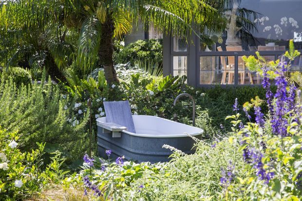 A freestanding outdoor bathtub is set among dense, lush planting beside a dark-framed glazed building. Tropical palms, flowering shrubs and purple flower spikes surround the tub, creating a secluded garden bathing area.