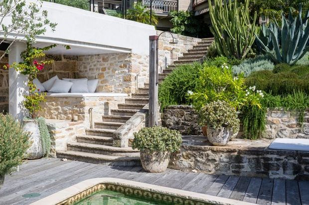 A sunlit landscaped pool terrace with a rustic stone retaining wall, built-in stone bench, broad timber decking, and a stepped stone stairway leading through dense Mediterranean-style planting. Large agaves, cacti, shrubs, and potted plants surround the outdoor space, with parts of a residential building visible above.