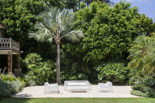 A lush tropical landscaped garden with a pale gravel or paved outdoor seating area, featuring a white outdoor sofa and two armchairs beneath mature palms and dense greenery. Part of a building with a decorative balcony is visible at the left edge.