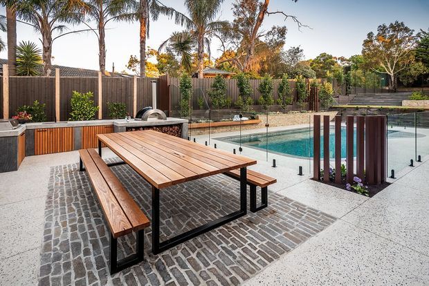 A landscaped outdoor entertaining area with a large timber dining table and benches on a dark brick-paved section beside a swimming pool. The pool is enclosed by frameless glass balustrading, with built-in outdoor cabinetry and barbecue facilities along the timber privacy fence. Vertical timber screening and mature palms add privacy and a resort-like atmosphere.