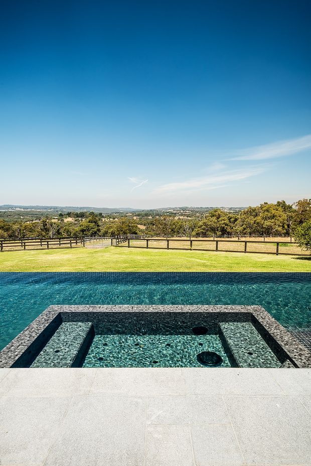 A large outdoor swimming pool with an integrated square spa or shallow lounging section, finished in dark patterned tile and surrounded by pale stone or concrete paving. Beyond the pool is a broad manicured lawn, timber post-and-rail fencing, mature trees and an expansive rural landscape under a clear blue sky.