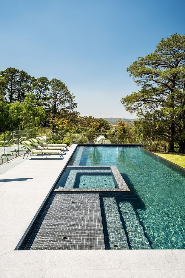 A bright outdoor pool area featuring a long rectangular swimming pool with integrated tiled steps and a raised square spa. Pale stone or concrete paving surrounds the pool, with lounge chairs on one side, glass pool fencing, and a leafy suburban landscape beyond.