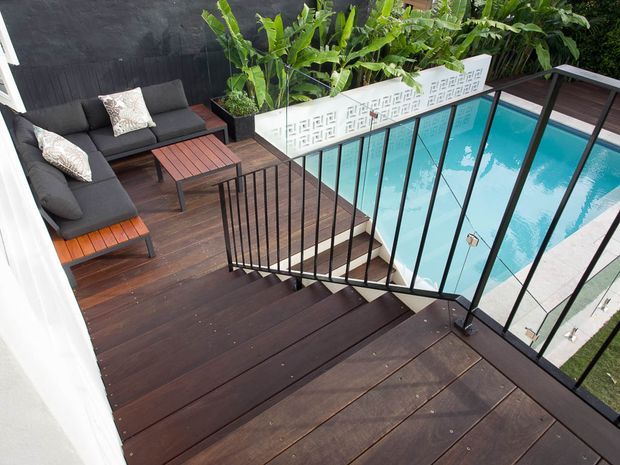 Elevated view of a dark timber deck and stairway with black metal balustrading, overlooking a bright rectangular swimming pool. A compact outdoor lounge with charcoal seating and timber coffee table sits beside lush tropical planting and a decorative breeze-block privacy wall.