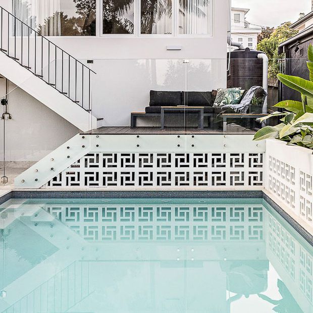A bright contemporary courtyard pool area with a turquoise swimming pool, patterned breeze-block privacy walls, white rendered surfaces, glass balustrades, and an elevated outdoor lounge with a dark sofa. A staircase connects the upper terrace to the pool level, while tropical planting borders the space.