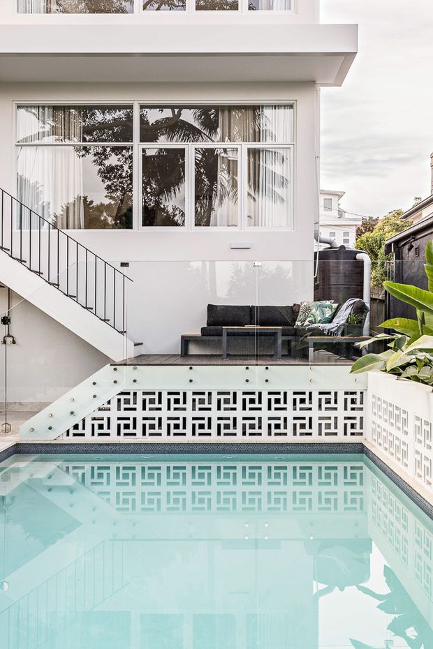 A compact urban courtyard pool beside a white multi-level residence. The image features a pale blue swimming pool, patterned breeze-block privacy walls, a glass balustrade, an external stair with slim black railing, and a small outdoor lounge with dark seating and tropical planting. Large white-framed windows reflect surrounding trees, creating a bright contemporary outdoor setting.
