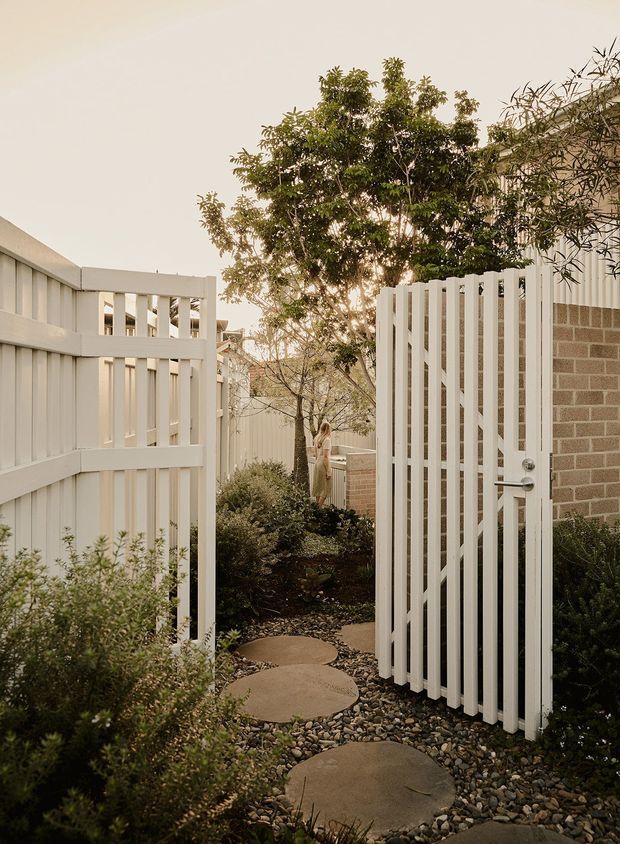 A landscaped garden passage viewed through an open white slatted gate and matching fence. Large circular stepping stones run through dark river pebbles and low planting toward a leafy courtyard, with a pale brick wall and additional screening beyond. The scene has a warm, softly lit contemporary garden character.