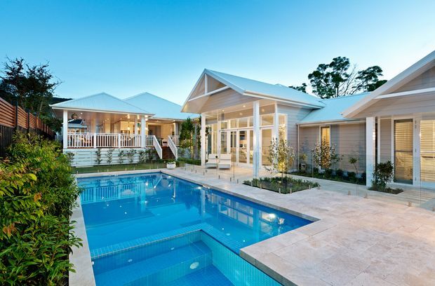A bright residential backyard centered on a large blue swimming pool with a shallow tiled section and surrounding pale stone paving. The pool is enclosed by a low, light-coloured single-storey home with multiple gabled roof forms, extensive glazed doors and windows, covered verandah areas, white posts and railings, and light grey weatherboard-style walls. Landscaped planting and timber screening frame the space.