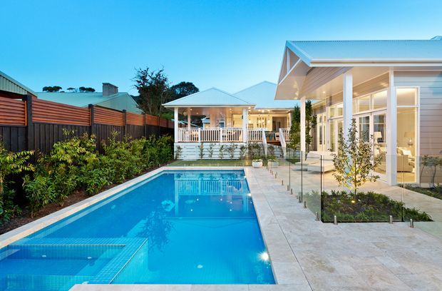 Bright exterior view of a contemporary single-storey home with a long rectangular swimming pool, covered entertaining pavilion, glazed living areas, pale rendered or weatherboard-style walls, and a light metal gabled roof. The pool terrace is paved in pale stone, with glass pool fencing and landscaped garden beds along a timber privacy fence.