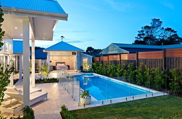 A landscaped residential backyard with a rectangular swimming pool enclosed by frameless glass fencing. The pool terrace includes pale tiled paving, lounge furniture, planting, and a small pavilion or pool house with a bright blue metal hipped roof. The main house and additional outbuildings feature white finishes and blue metal roofing, while a timber privacy fence lines the boundary. The image is photographed at dusk with warm interior lighting and a vivid blue sky.