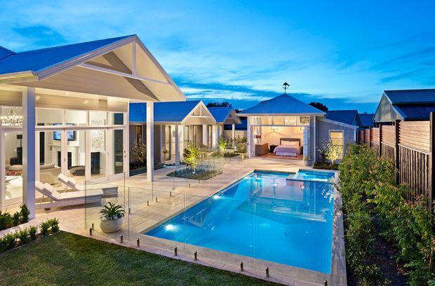 A professionally landscaped residential courtyard viewed at dusk, centred on a bright rectangular swimming pool with surrounding paved terraces. Several connected pavilion-like buildings feature pitched gable roofs, light-coloured rendered walls, extensive glazed doors and windows, and warm interior lighting. Outdoor seating, planting, privacy fencing and poolside landscaping create a resort-like backyard setting.