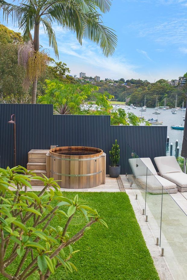 A landscaped outdoor terrace overlooking a sheltered marina, with a round timber hot tub on a small timber deck, dark vertical privacy fencing, glass balustrades, white reclining loungers, tropical planting, lawn, and a potted plant. The scene is bright and leafy with boats and wooded hills visible beyond the property.