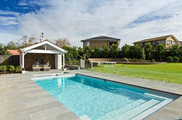 A landscaped backyard with a large rectangular swimming pool, broad grey tiled paving, and frameless glass pool fencing. A small white gabled pool pavilion with an outdoor seating area sits beside the pool, with manicured lawn, hedges, and neighbouring suburban houses beyond.