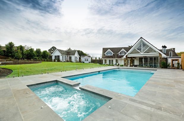 A spacious landscaped residential property viewed from the pool terrace, featuring a large swimming pool and attached bubbling spa in the foreground. Beyond the water is a broad lawn and a substantial light-coloured gabled house with prominent glazed triangular facade, multiple dormer windows, and an adjoining glazed pool-house or entertaining pavilion. Paved stone terrace surrounds the pool, with mature planting and additional outbuildings in the background.
