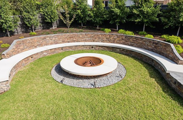 A landscaped outdoor courtyard featuring a circular fire pit with a pale stone or concrete surround, set within a neatly mown lawn and encircled by a curved built-in stone retaining wall with integrated white seating. Dense planting and privacy fencing frame the space.