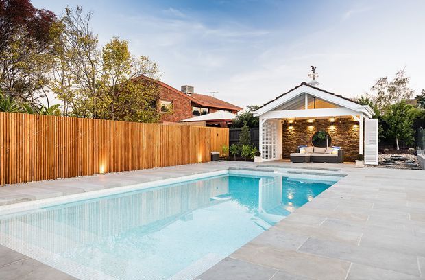 A landscaped backyard with a large rectangular swimming pool surrounded by pale stone paving. At the far end is a detached white gabled pool pavilion or outdoor lounge with timber-clad feature walls, built-in seating, warm ceiling lights, and an outdoor fireplace or television wall. Timber privacy fencing, mature planting, and a brick house are visible beyond.