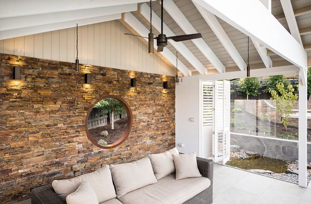 Bright covered outdoor lounge with a large cushioned sofa, white-painted exposed rafters and beams, a dark ceiling fan, and a striking stacked-stone feature wall with a circular window. The space opens to a landscaped courtyard through white louvred screens and glazing.