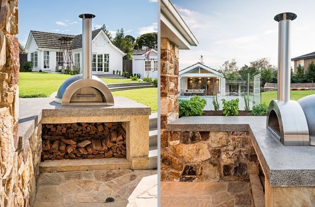 Two views of a built-in outdoor pizza oven and firewood storage set into a rustic stone masonry structure. The oven has a smooth metallic dome and tall flue, with a speckled stone or concrete benchtop above a stacked firewood recess. The setting includes a landscaped residential courtyard and a white gabled house visible beyond.