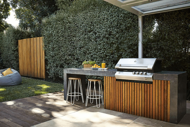 A landscaped outdoor entertaining area with a built-in barbecue kitchen, timber-slatted cabinetry, dark stone benchtop, bar stools, and a covered overhead structure. The kitchen sits beside a timber deck and lush screening vegetation, with a freestanding timber privacy screen and casual outdoor seating nearby.