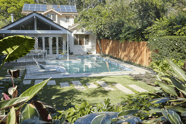 A landscaped residential backyard with a rectangular swimming pool, paved pool terrace, lawn and lush tropical planting. The house has a light-coloured exterior with a dark gabled roof, extensive glazed doors, and visible rooftop solar panels. Timber fencing encloses the garden.