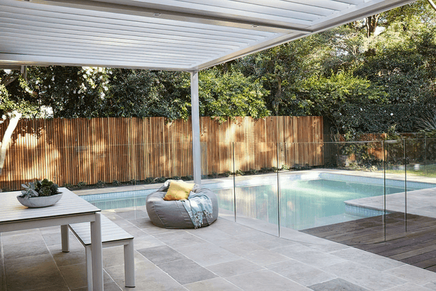 A bright, contemporary outdoor entertaining area beside a swimming pool. The space features a large white louvred pergola, pale stone paving, a timber dining table with bench seating, a beanbag lounge chair, frameless glass pool fencing, a timber privacy fence, and surrounding greenery.