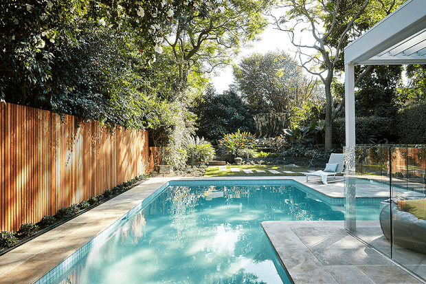 A sunlit residential backyard with a rectangular swimming pool, paved pool surround, timber privacy fencing, landscaped garden, and a covered outdoor area with glass screening and lounge seating. Mature trees and dense planting create a private, leafy setting.