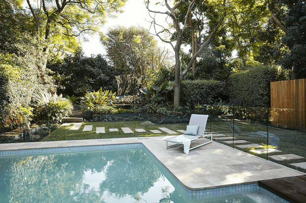 A tranquil landscaped backyard with a rectangular swimming pool, pale paved pool terrace, white reclining sun loungers, and a glass balustrade. Dense mature planting surrounds the space, with stepping stones across the lawn and a vertical timber privacy screen along one boundary.