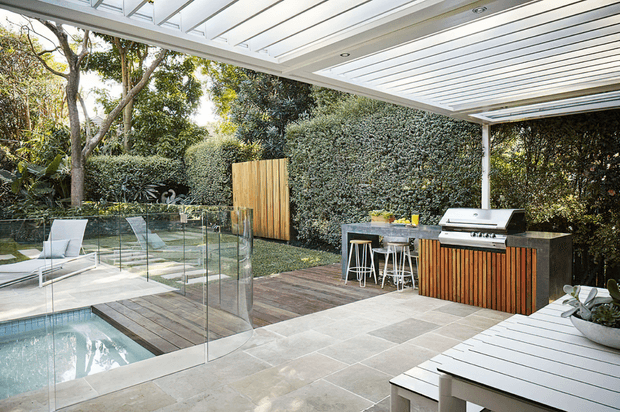 A bright, contemporary outdoor entertaining area beside a swimming pool, featuring a covered timber deck, built-in outdoor kitchen with barbecue and bar seating, glass pool fencing, pale stone paving, and lush landscaped screening.