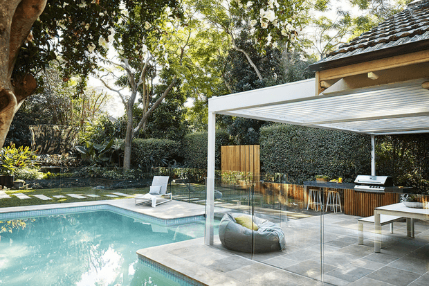 A landscaped residential outdoor entertaining area with a turquoise swimming pool, large tiled terrace, frameless glass pool fencing, and a covered outdoor kitchen and dining zone. The contemporary pergola has a white slatted roof, while timber cabinetry, a built-in barbecue, stools, outdoor seating, and a dining table create a polished alfresco setting surrounded by mature greenery.