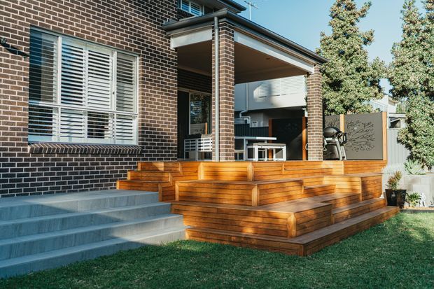 Exterior view of a contemporary brick house with a covered outdoor entertaining deck, broad timber steps, built-in timber seating, white plantation shutters, and a landscaped lawn. The raised deck includes an outdoor dining area and barbecue, with a privacy screen at the side.