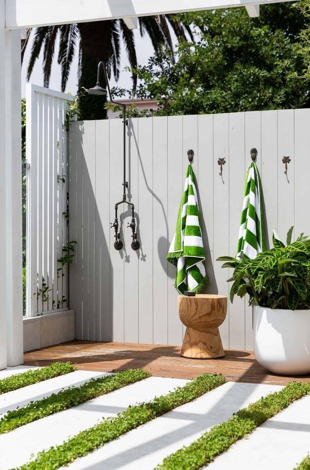Bright outdoor shower courtyard with a wall-mounted rain shower, white vertical-panel privacy screen, striped green-and-white towels, potted tropical plants, and a timber stool or planter. White stepping slabs with narrow strips of greenery lead through the foreground, creating a fresh resort-like outdoor setting.